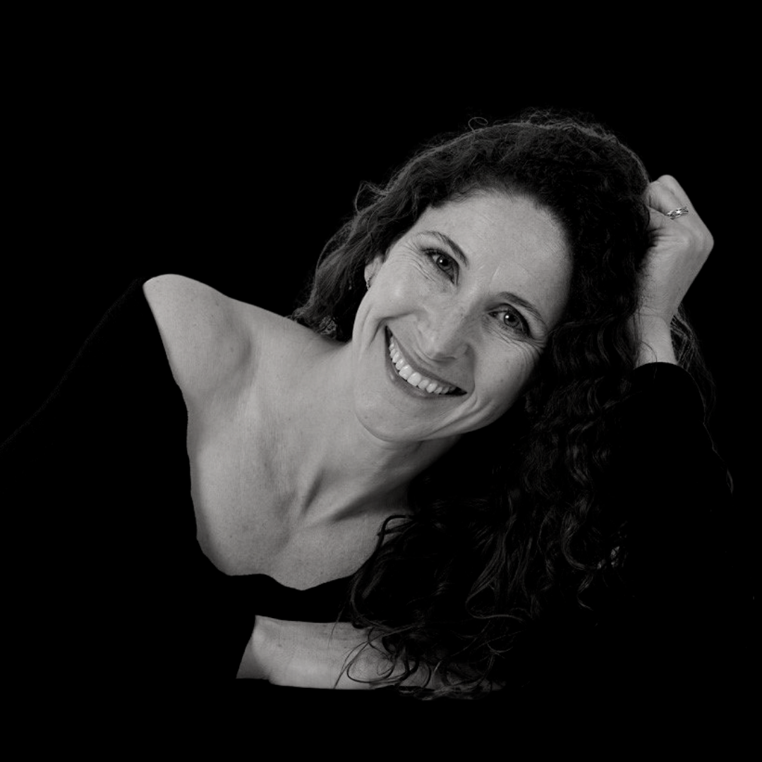 A black and white image of Carrie Thiel - Carrie is wearing a black top, leaning her head on her hand and is smiling.